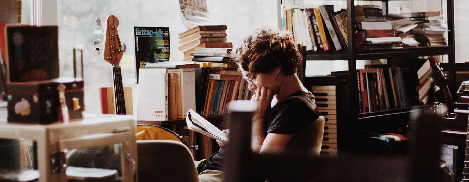 woman, surrounded by books, sits and reads in front of a picture window