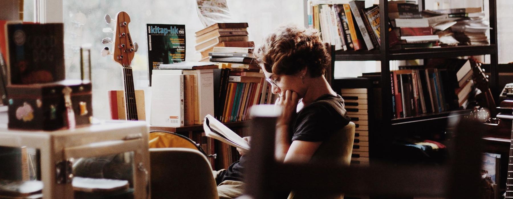 woman, surrounded by books, sits and reads in front of a picture window